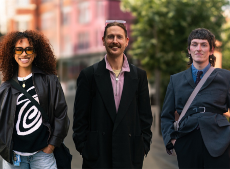 Three individuals are standing outdoors, smiling at the camera. The person on the left is female and has dark skin, she has curly hair, is wearing a leather jacket, graphic sweater, and sunglasses. The person in the middle has a mustache, is wearing a black coat over a light purple zip-up shirt, and has glasses on top of their head. The person on the right has short hair, is wearing a suit with a belt over the jacket, and has a crossbody bag. The background is a city street with blurred buildings and trees.