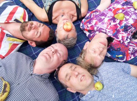 Five people lie on their backs in a circular arrangement on a blue plaid blanket. They appear relaxed, with eyes closed. The person in the center has a red apple placed over their mouth, while the others have fruits such as a banana and pears resting on their clothing. Their outfits are colorful and varied, adding to the playful and relaxed atmosphere of the scene.